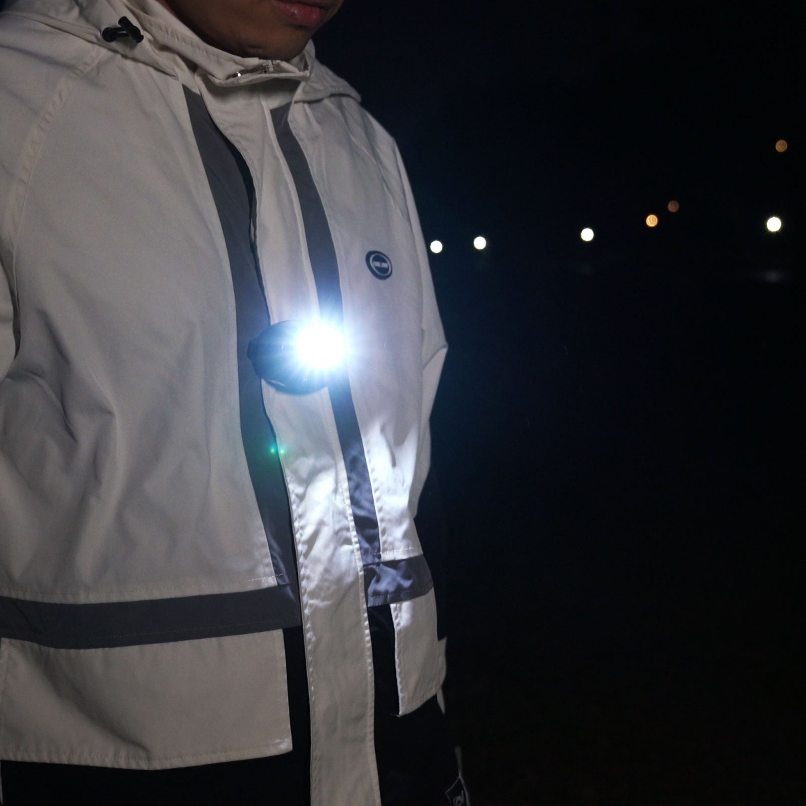 Image of Person wearing a reflective jacket with a light attached, standing against a dark background with blurred lights.